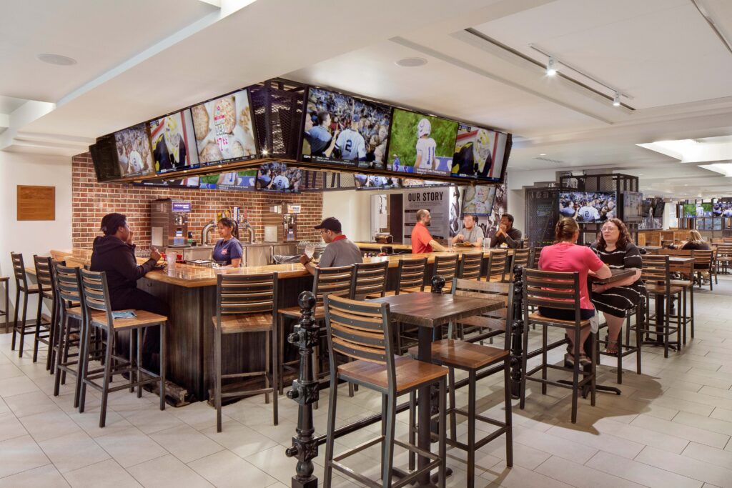 Interior of a sports bar with patrons seated at a U-shaped wooden bar under a central ring of television screens showing sports games.