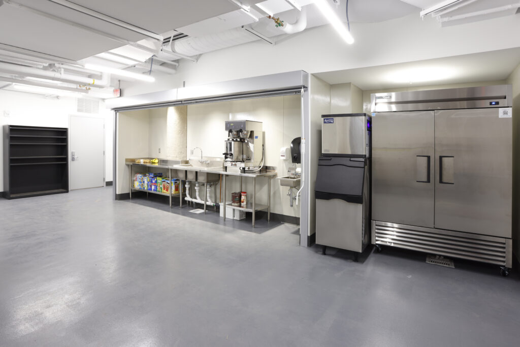 A commercial kitchen alcove with a stainless steel counter, sink, coffee machine, ice maker, and a two-door refrigerator.