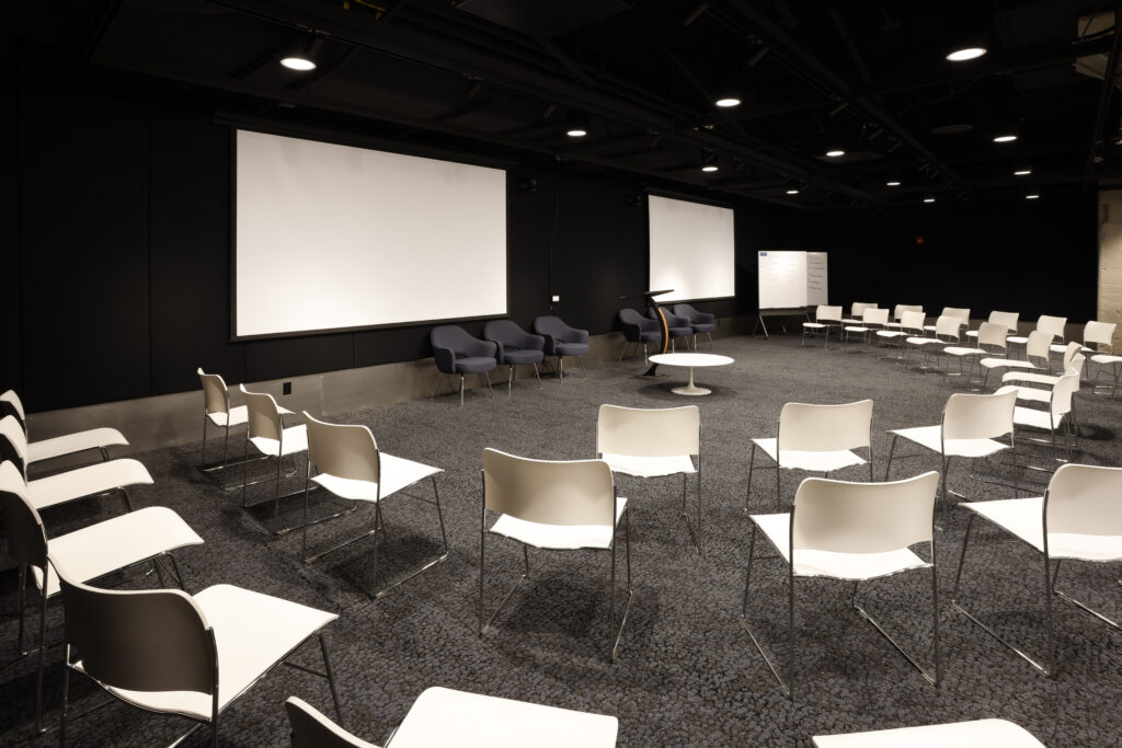 A seminar room with white chairs arranged in a circle, facing two large projector screens and a speaker’s podium.