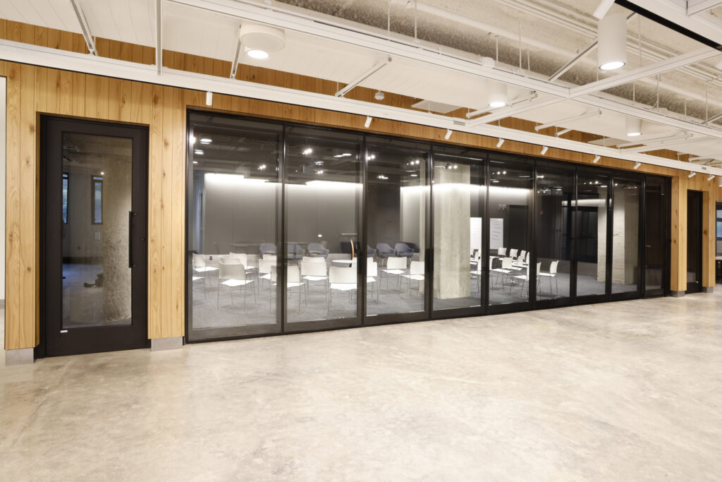 A modern meeting room enclosed by a floor-to-ceiling glass wall, featuring rows of chairs arranged in a large, open space.