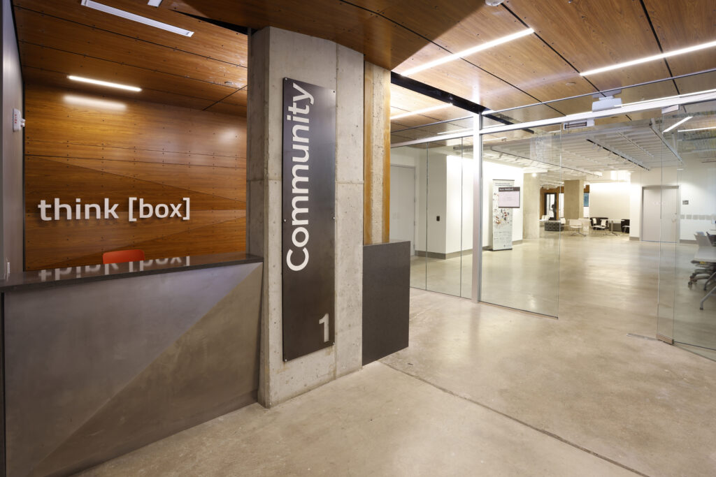 Reception area featuring a wood-paneled wall with the text “think [box],” a concrete pillar labeled “Community 1,” and glass doors leading to a bright, open office space.