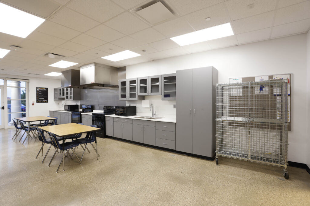 A kitchen area featuring grey cabinetry, stainless steel appliances, two dining tables with chairs, and a metal storage cart.