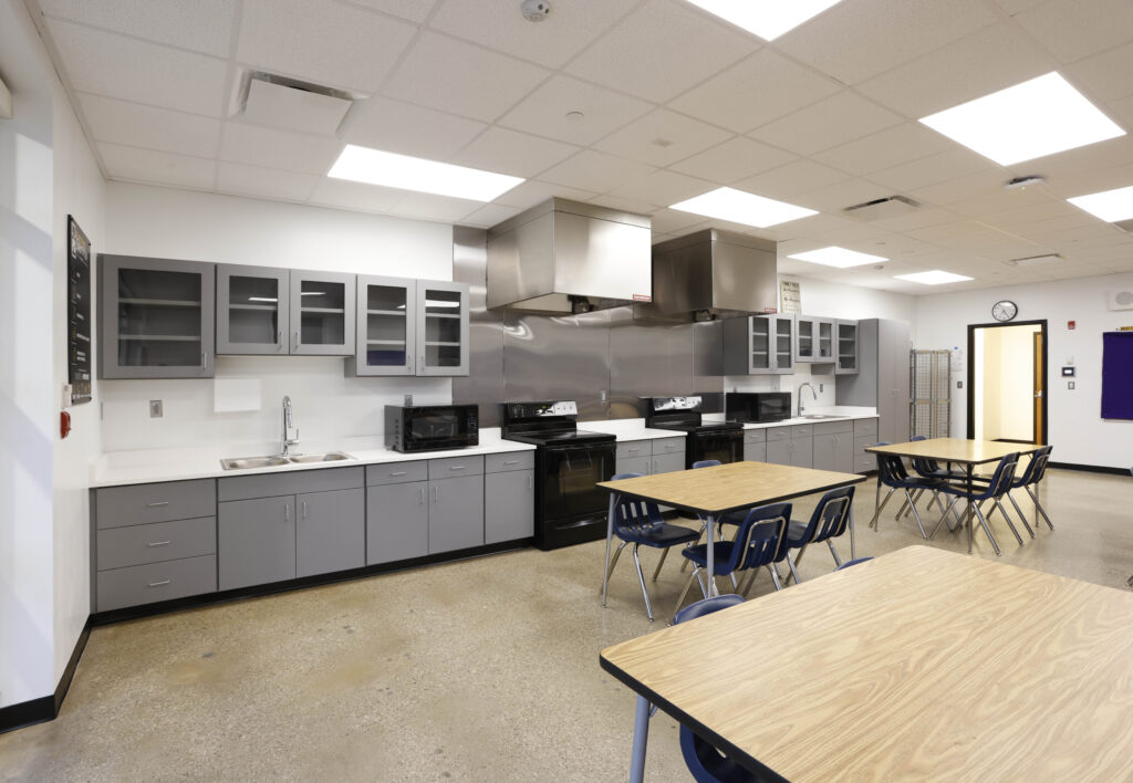 A classroom kitchen featuring gray cabinets, stainless steel appliances, and several tables with blue chairs.