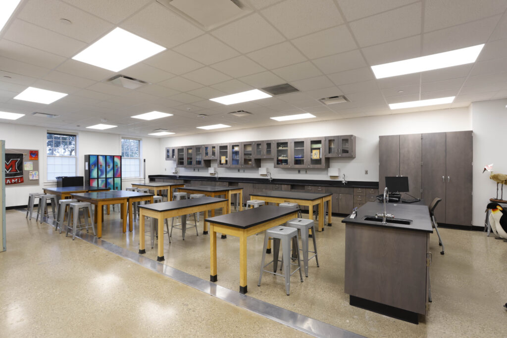 A bright science classroom with multiple lab tables, stools, perimeter cabinets, and a sink station.