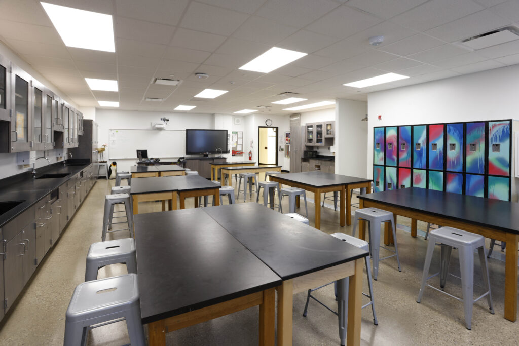 A high school science classroom with rows of lab tables, stools, a whiteboard, a large monitor, and a wall of colorful graffiti-style lockers.
