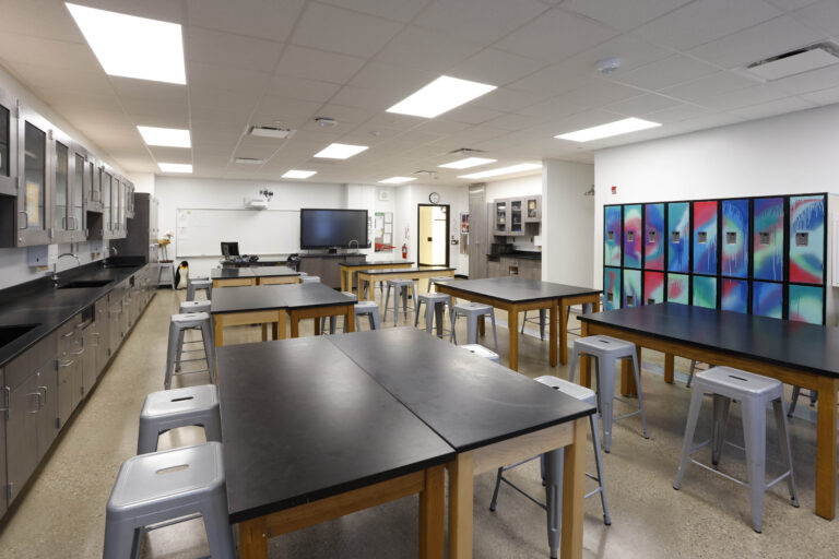 A high school science classroom with rows of lab tables, stools, a whiteboard, a large monitor, and a wall of colorful graffiti-style lockers.