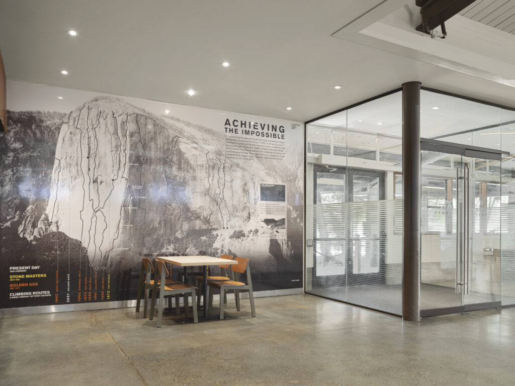 A small table and four chairs sit in front of a wall mural depicting El Capitan with climbing route lines and the text “Achieving the Impossible,” next to a glass-walled conference room.