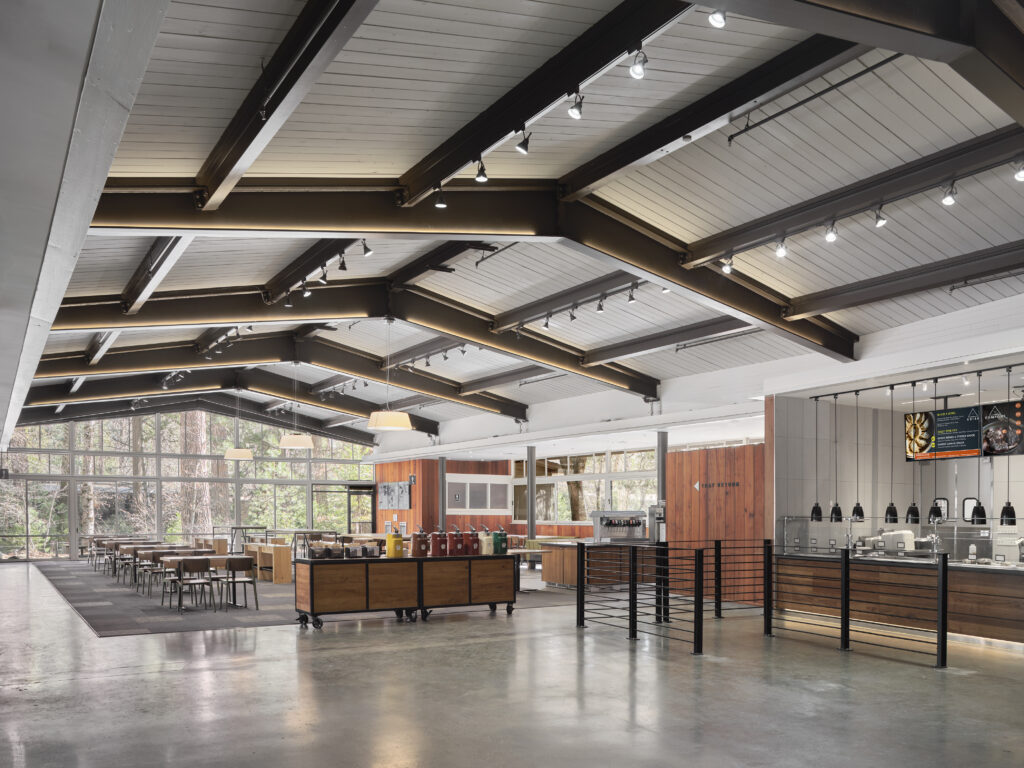 Spacious cafeteria with wooden ceiling beams, dining tables, a self-service beverage station, and a food service counter.