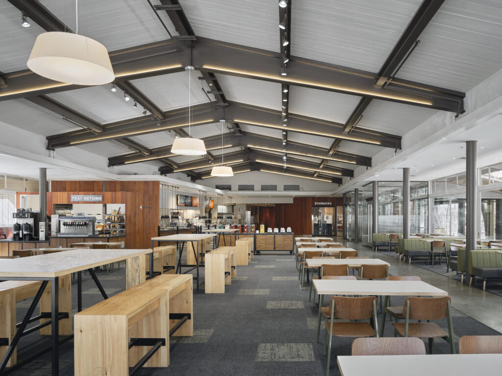Interior view of a modern dining area with wooden benches, tables, chairs, and a Starbucks counter in the background.