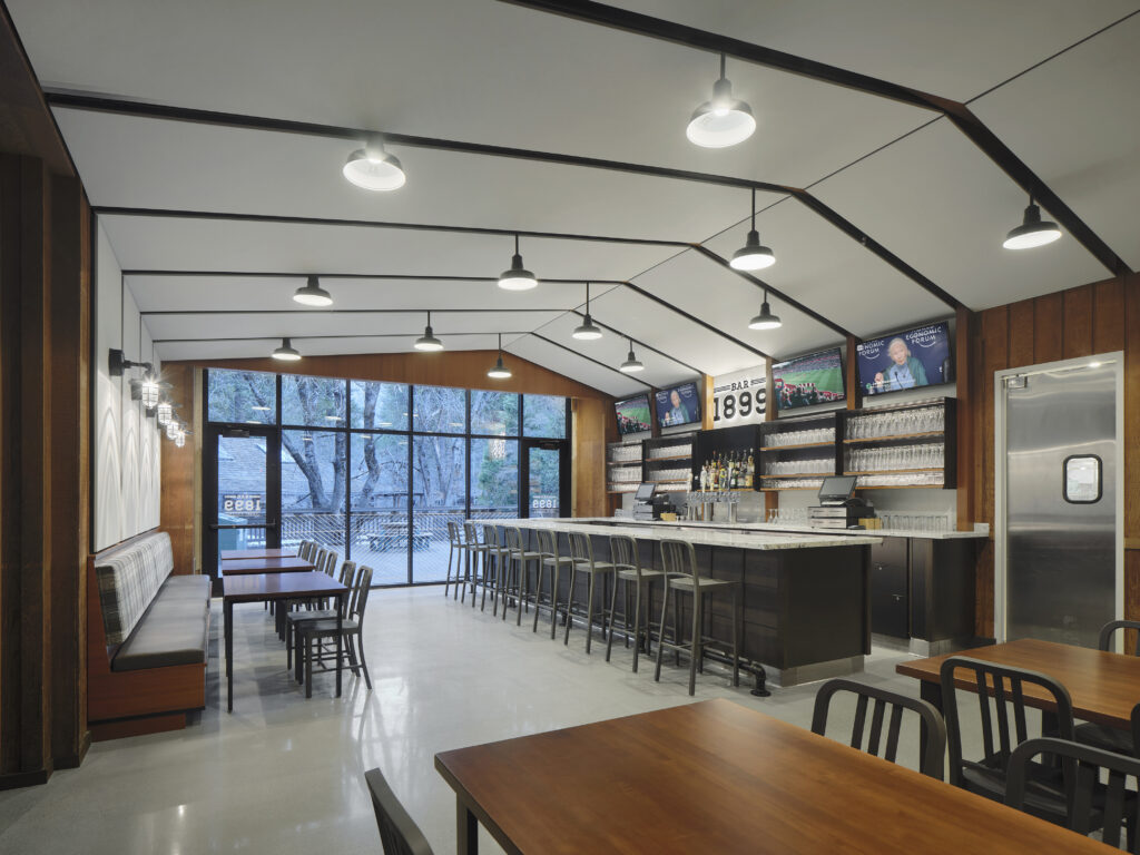 Interior view of a bar and restaurant featuring a long marble-topped counter with stools, wooden tables, and large windows looking out to trees.