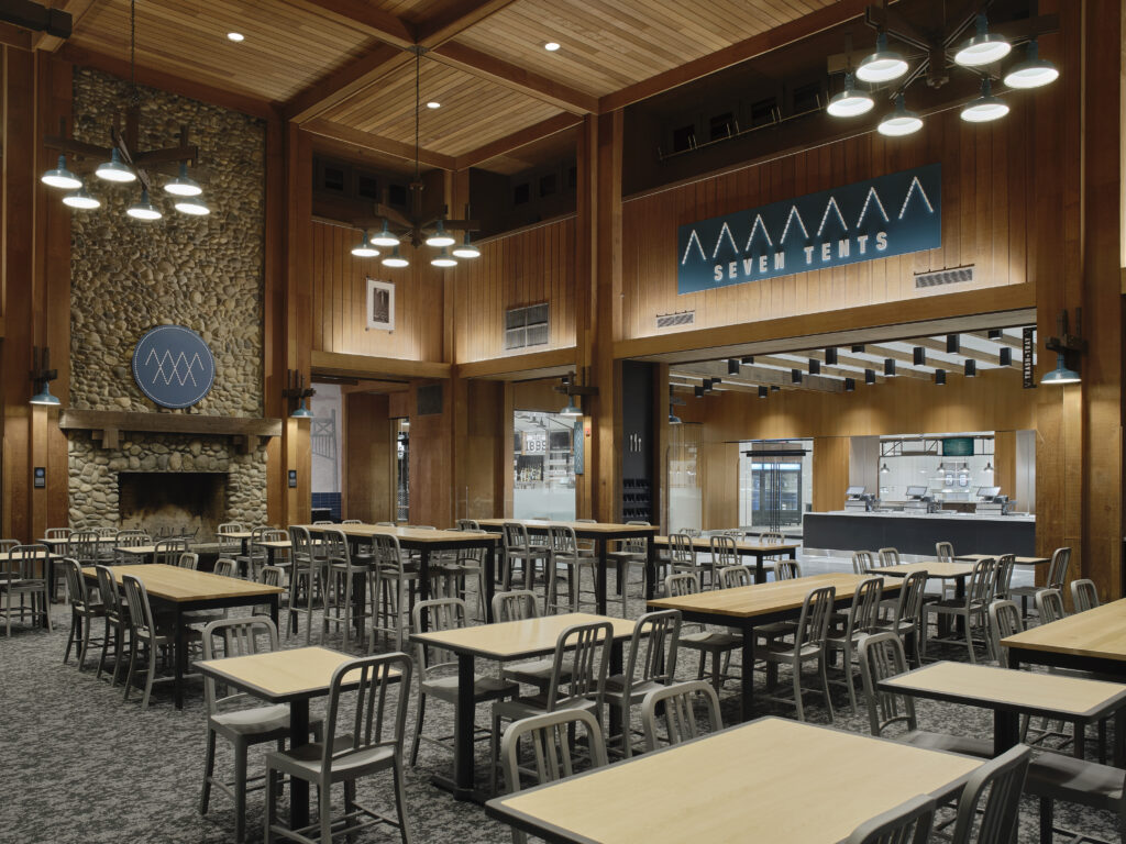 Dining hall with many wooden tables, metal chairs, a stone fireplace, and a large sign that reads “SEVEN TENTS.”