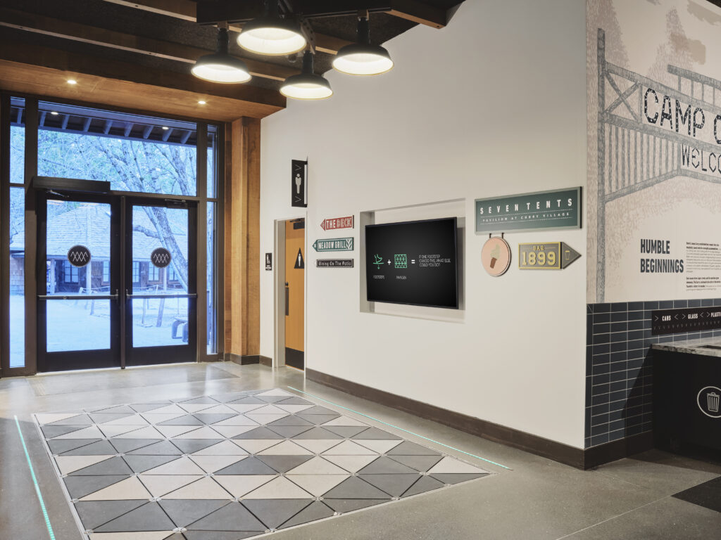 Interior lobby of Curry Village featuring a digital screen, directional signage, and a glass door entrance looking out to trees.