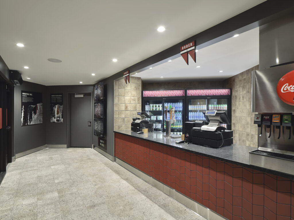 A concession stand featuring a dark countertop, a red tiled front, two point-of-sale terminals, a drink fountain, and a refrigerated display case with an “Order” sign above.