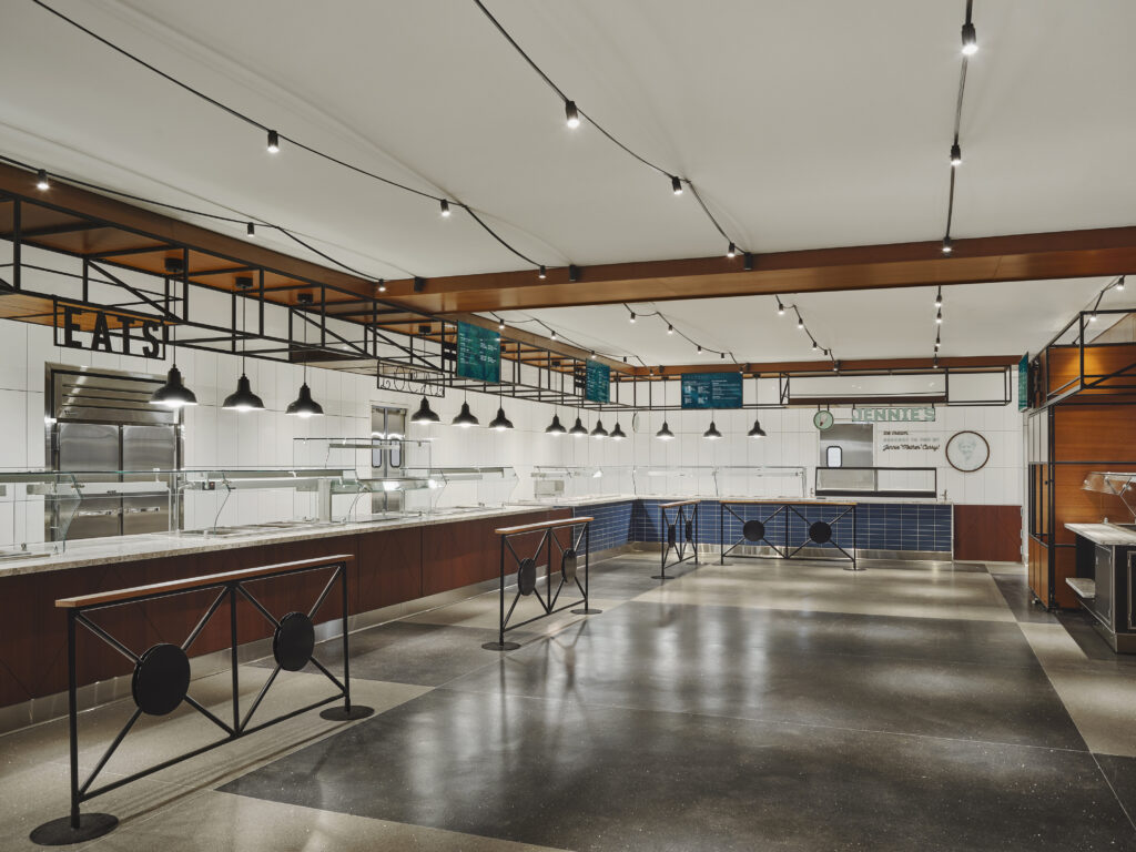 Empty school-style cafeteria serving line with blue tiled walls, hanging menu boards, and black pendant lights.
