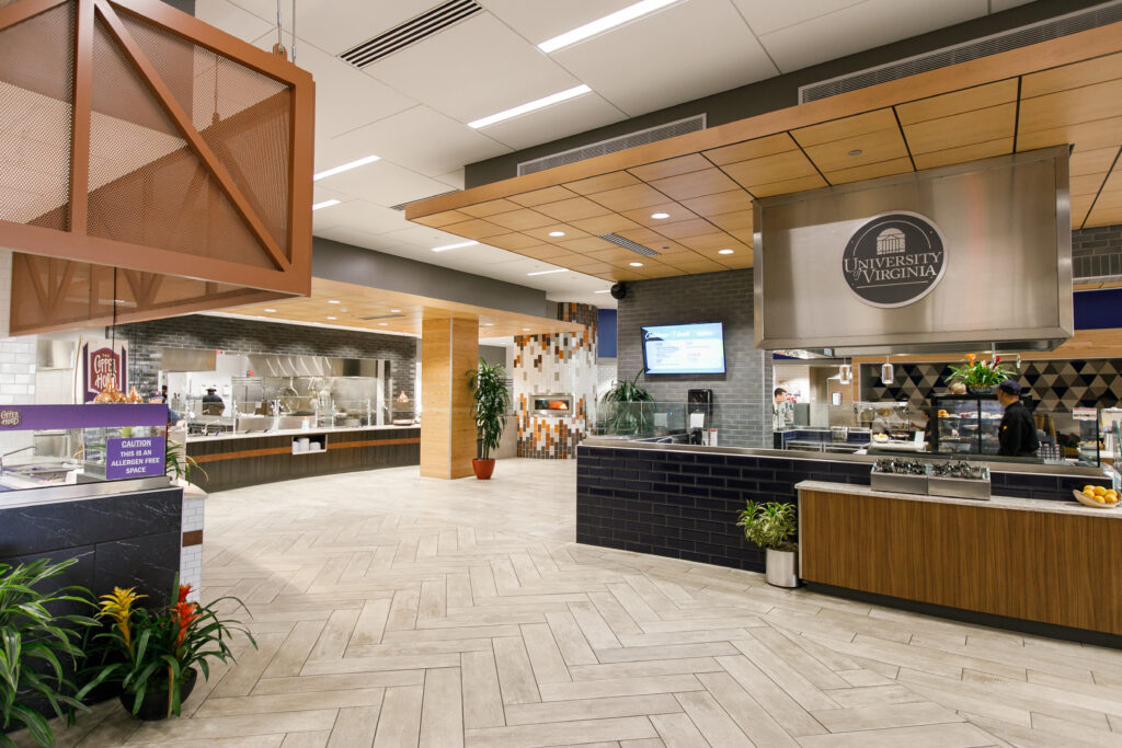 An interior view of a modern university dining hall featuring serving stations, a University of Virginia logo on a stainless steel hood, and a chevron-patterned floor.
