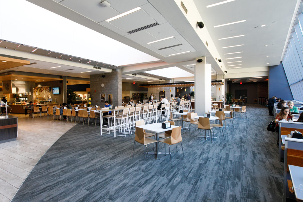 A large, modern university dining hall with numerous tables and chairs, a cafeteria service area, and students eating.