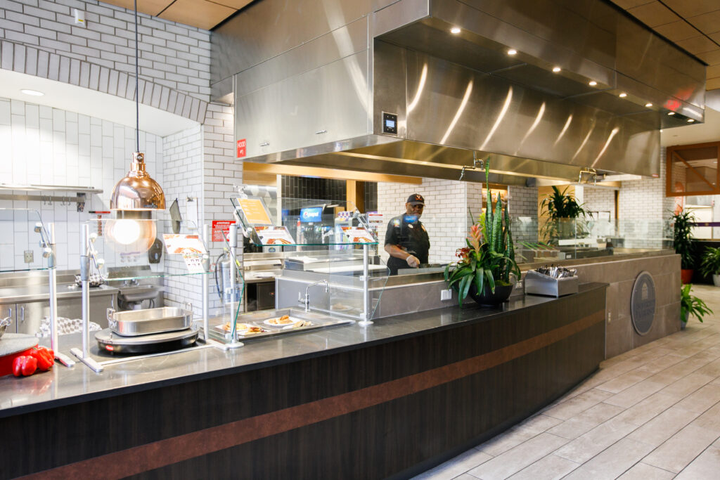 A food service worker stands behind a long stainless-steel cafeteria counter with a food display case.