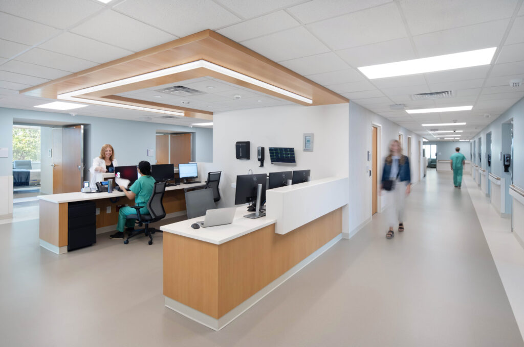 A hospital nursing station with staff members at desks, and people walking in the adjacent hallway.