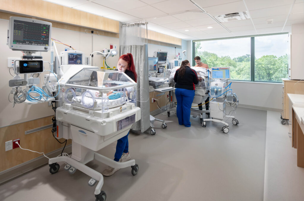 Two neonatal nurses care for infants in incubators inside a hospital nursery.
