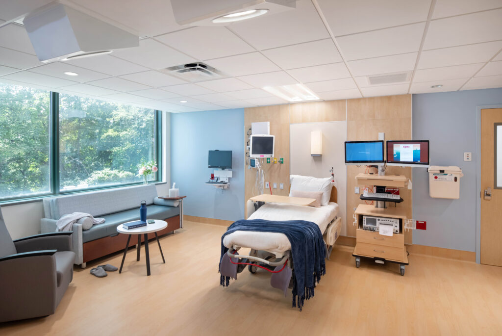 A hospital room with a patient bed, medical monitors on a rolling cart, a sofa, and a large window.