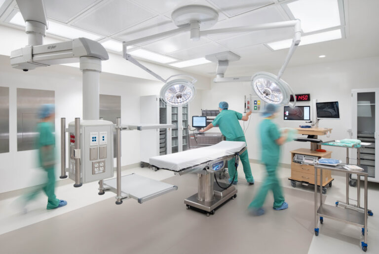 Medical staff in scrubs working in a modern operating room with an examination table and overhead surgical lights.