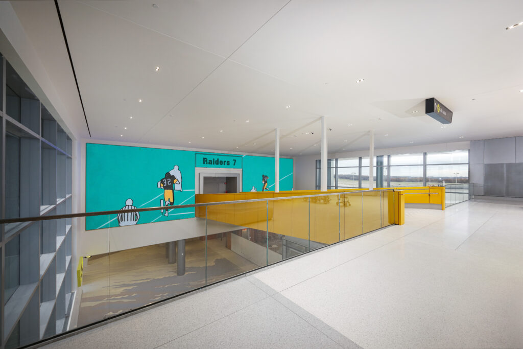 An indoor walkway with a yellow glass railing overlooking a lower level, featuring a large teal mural of a football player labeled “Raiders 7.”