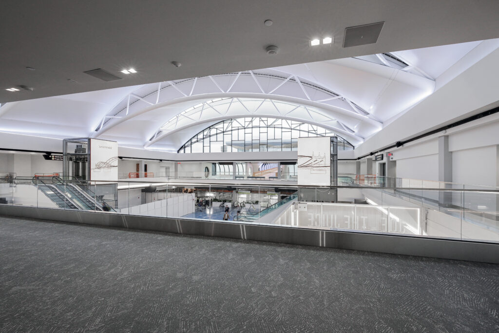 A modern, multi-level airport terminal with a vaulted ceiling, glass railings, and escalators connecting the floors.
