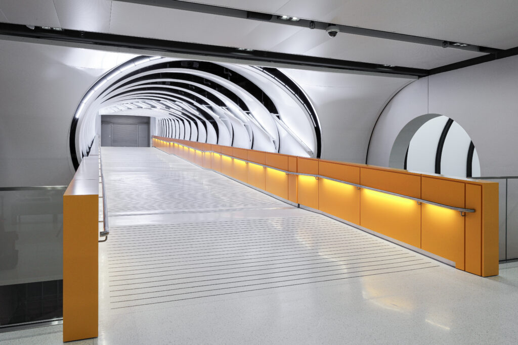 An enclosed pedestrian bridge with white arched ceiling ribs, a white floor, and a long yellow guardrail with integrated lighting.