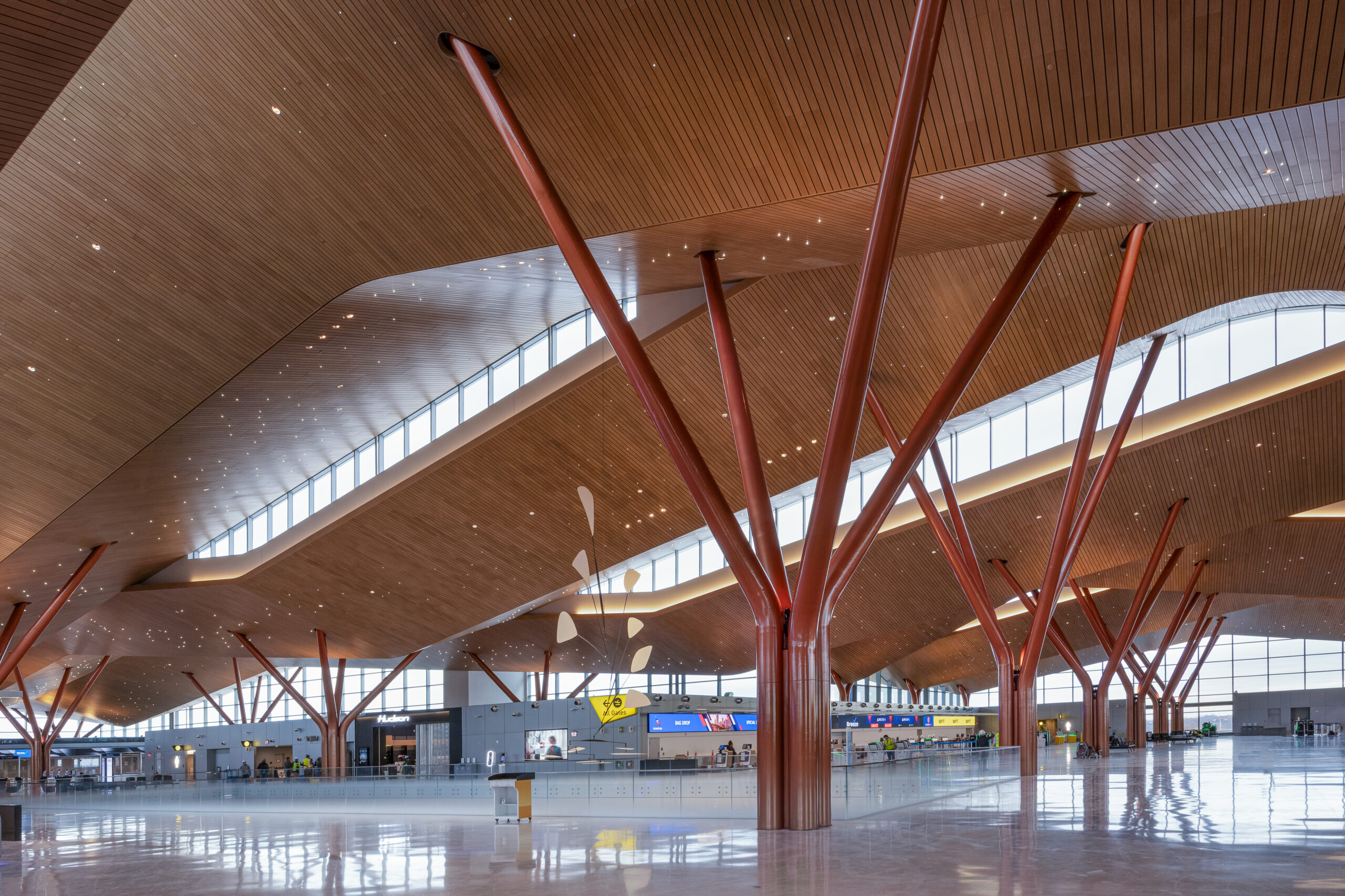 Modern airport terminal interior with a high, slatted wood ceiling supported by large, red branched columns and a polished floor.