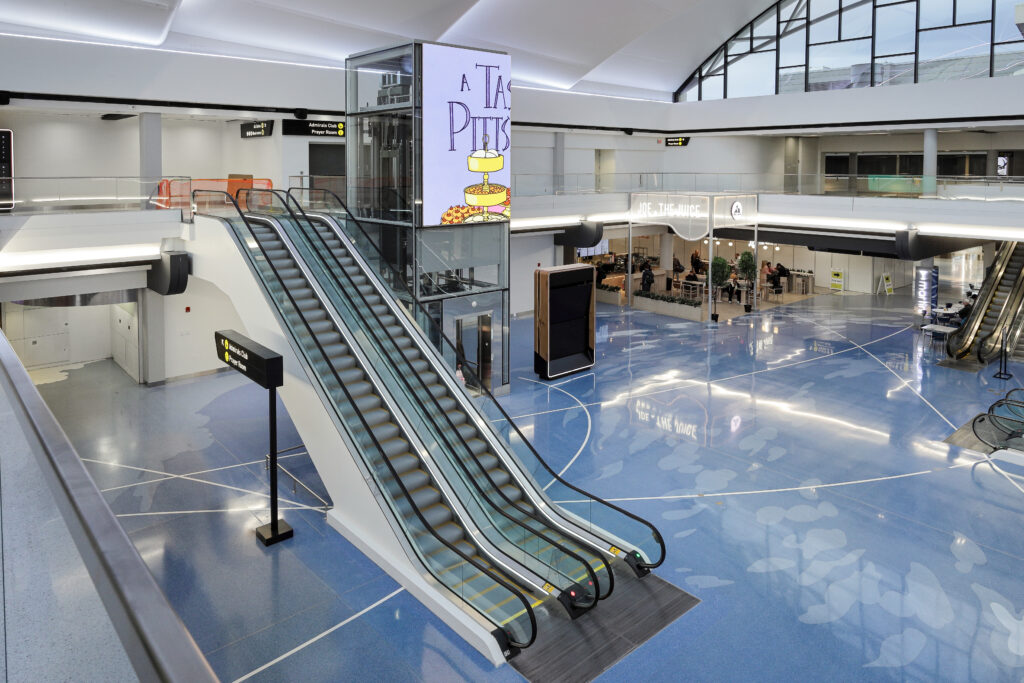 Elevated view of an airport terminal featuring a central glass elevator, a large escalator, and a Joe & The Juice cafe on the blue floor.