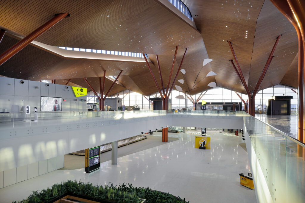 An interior view of a spacious, modern airport terminal with a high, wood-paneled ceiling, glass railings, and airport signage.