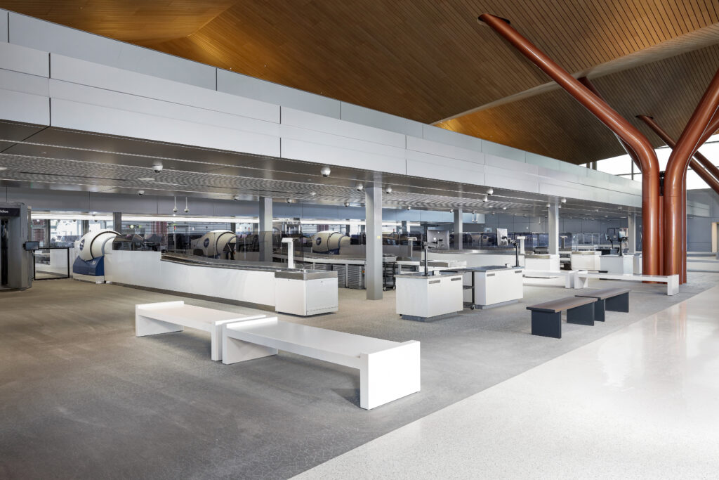 Empty airport security screening area with white check-in counters, metal benches, and overhead structural supports.