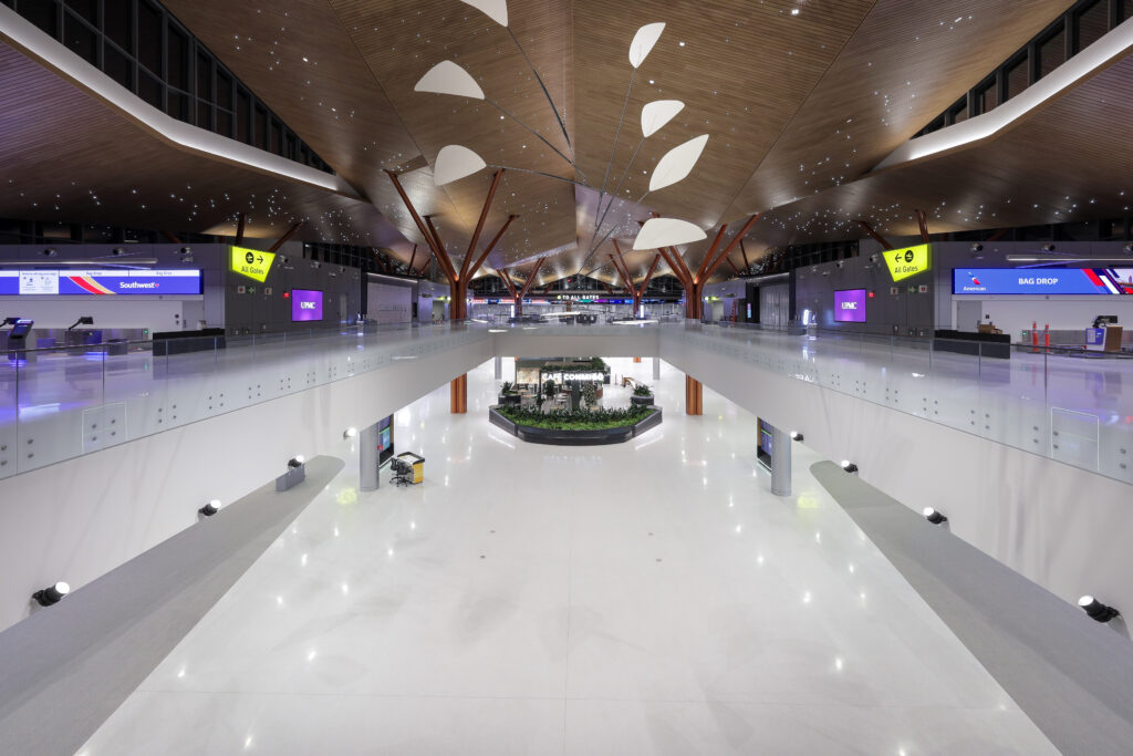 Interior view of a modern, spacious airport terminal with white floors, high wood-paneled ceilings, gate check-in counters, and a central retail kiosk.