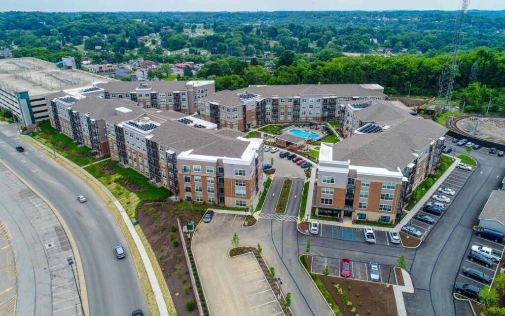 Aerial view of a large multi-story apartment complex with a central swimming pool, parking lots, and surrounding wooded hills.