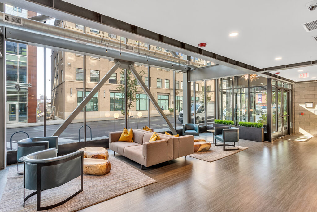 Modern lobby seating area with a sofa, lounge chairs, and wooden stump tables in front of large windows overlooking a city street.