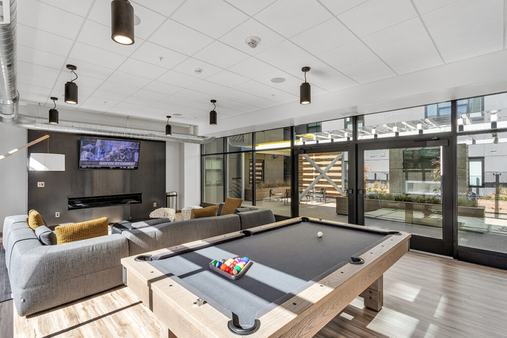 A lounge area with a grey sectional sofa, a wall-mounted TV, and a pool table in the foreground.