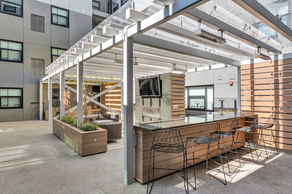 Outdoor apartment patio featuring a pergola, a stone wall with a mounted television and fireplace, and a bar counter with four wire stools.