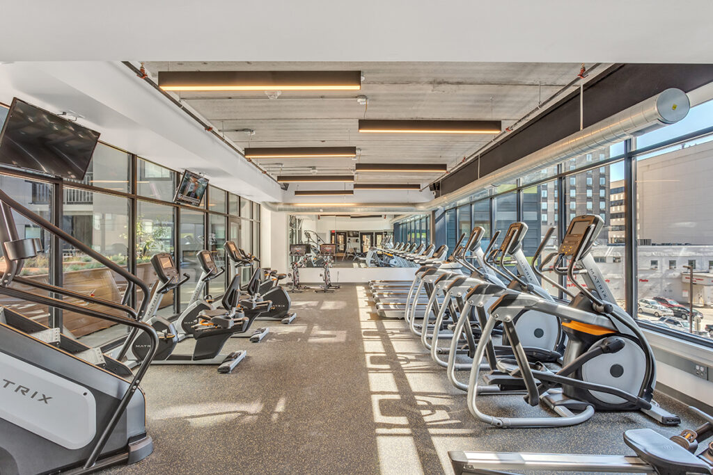 A gym with rows of Matrix elliptical machines and treadmills facing floor-to-ceiling windows.