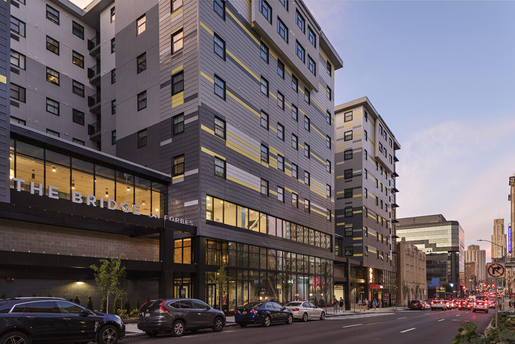 The Bridge on Forbes apartment building at dusk, featuring a multi-story facade with gray and yellow accents and ground-floor retail along a city street.