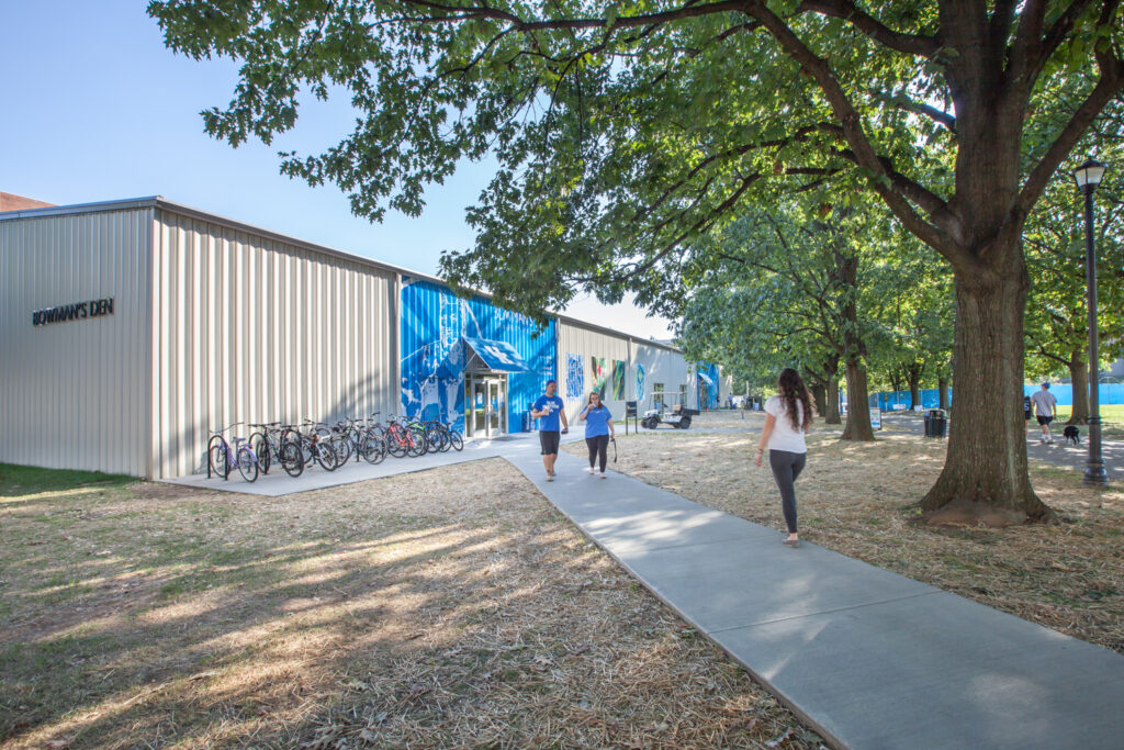 People walking on a sidewalk toward the entrance of a building labeled “Bowman’s Den,” with a row of bicycles parked outside.