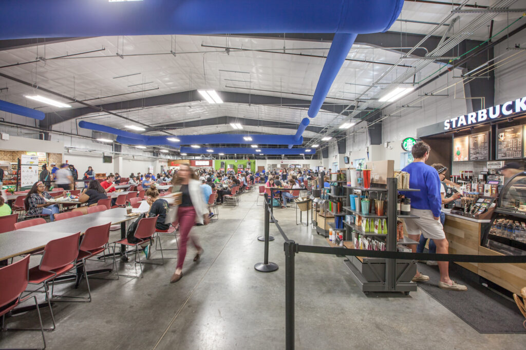 A busy university dining hall with a Starbucks counter on the right and rows of tables filled with students.