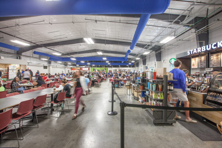 A busy university dining hall with a Starbucks counter on the right and rows of tables filled with students.