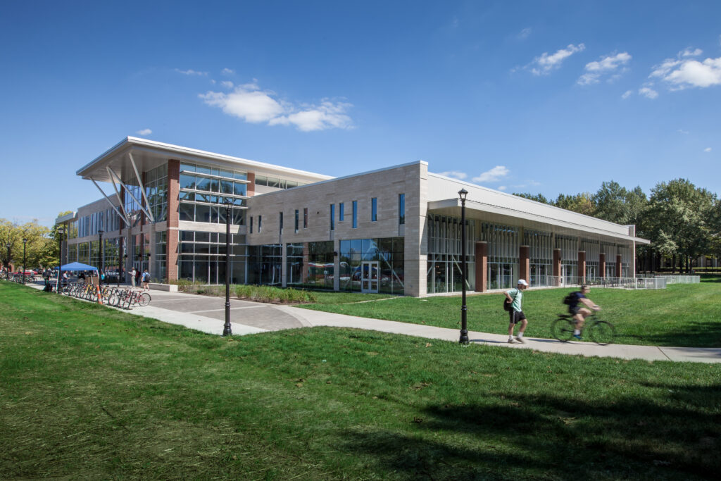 A modern multi-story building with a glass and stone facade, set on a campus lawn with people walking and cycling on a path.