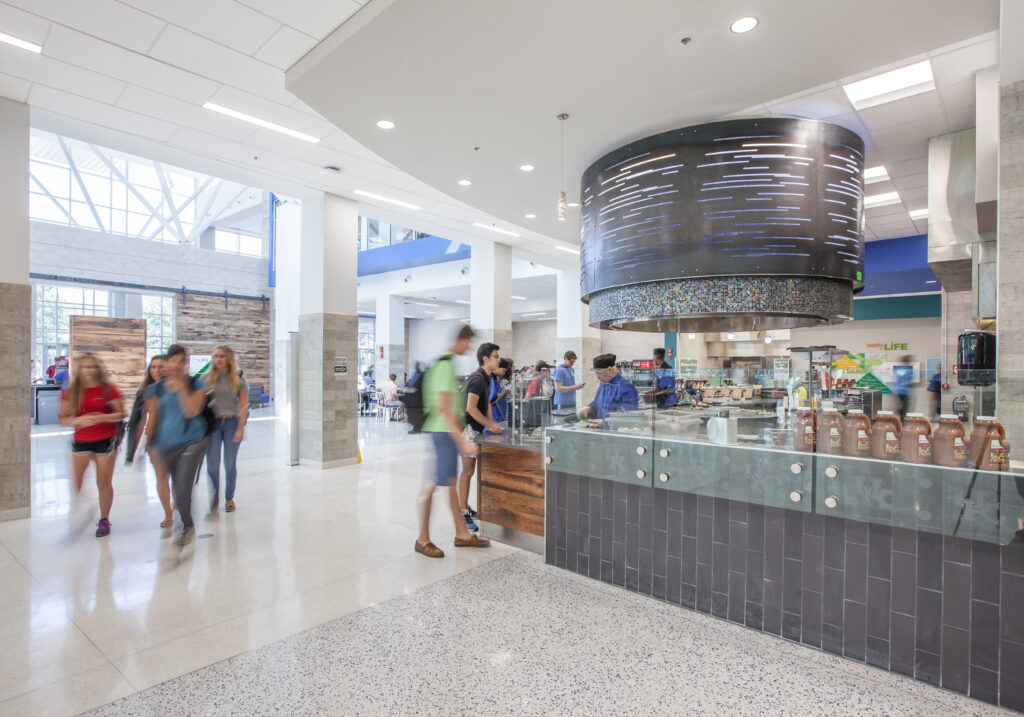 Students walk through a bright, modern campus food court with a circular service counter and prep area.