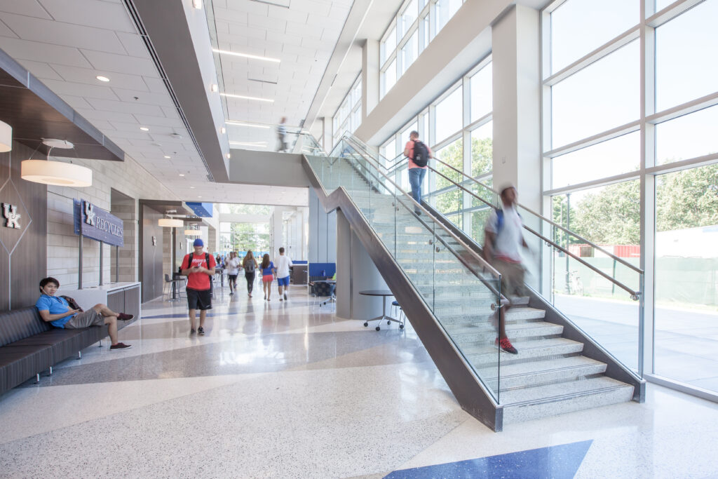 Students walking through a bright, multi-level building lobby with a central glass staircase.