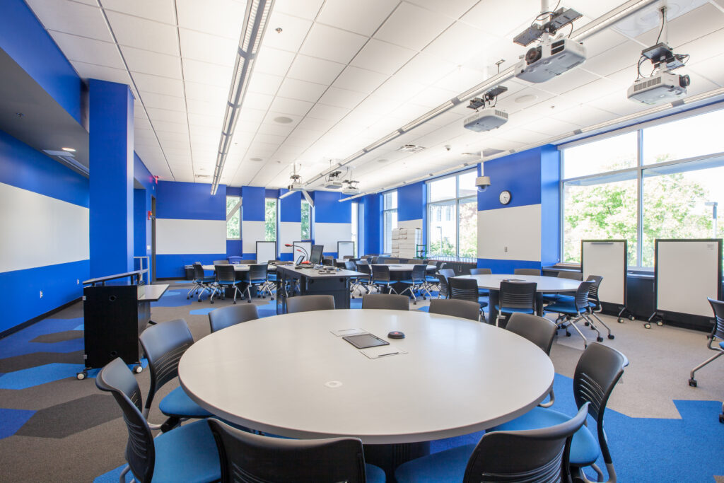 A modern classroom with several round tables and blue chairs, featuring blue walls, large windows, and ceiling-mounted projectors.