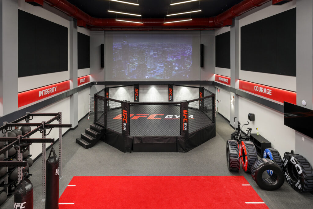 A UFC gym training area with a central octagon cage, a large projection screen displaying a city skyline, and surrounding fitness equipment.
