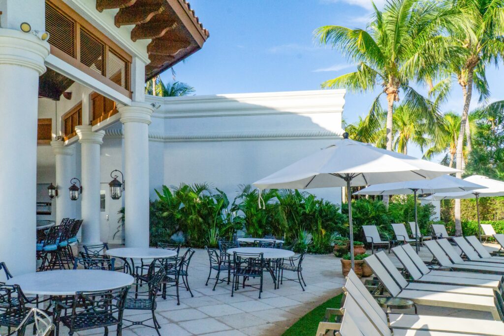 A resort patio with round dining tables, chairs, and rows of lounge chairs under umbrellas, surrounded by palm trees.