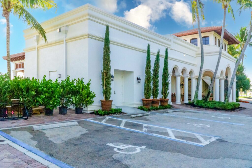 White building with arched columns, potted cypress trees, and a handicap-accessible parking space in the foreground.