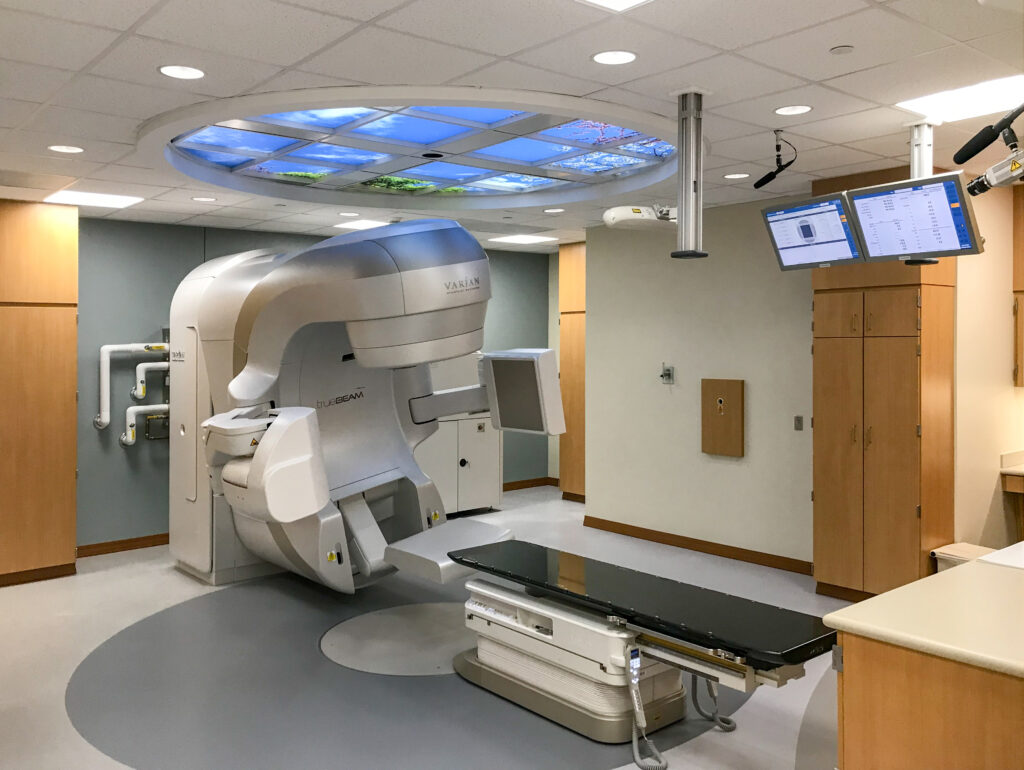 A Varian TrueBeam linear accelerator radiotherapy machine with an adjustable treatment table in a clinical room.